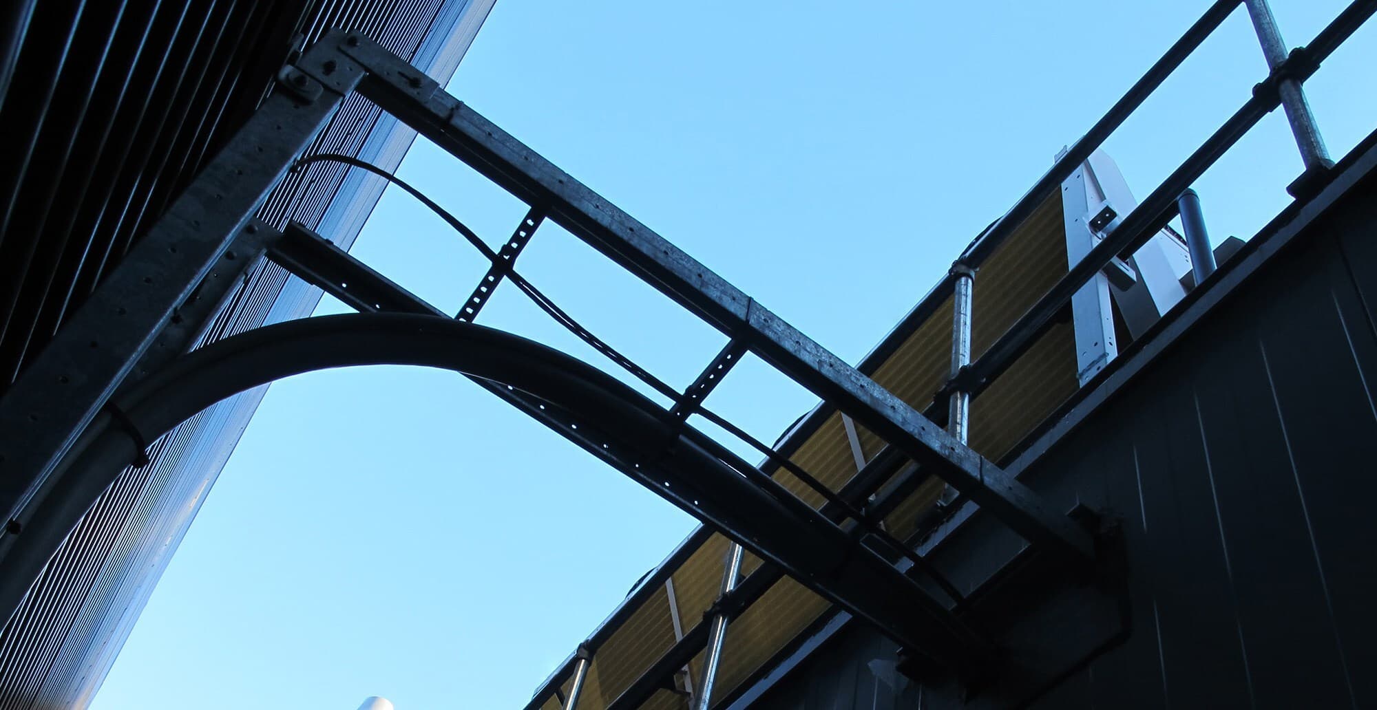 Low-angle view of metal framework, curved pipe, and railing against a blue sky, with yellow panel and exposed scaffolding.